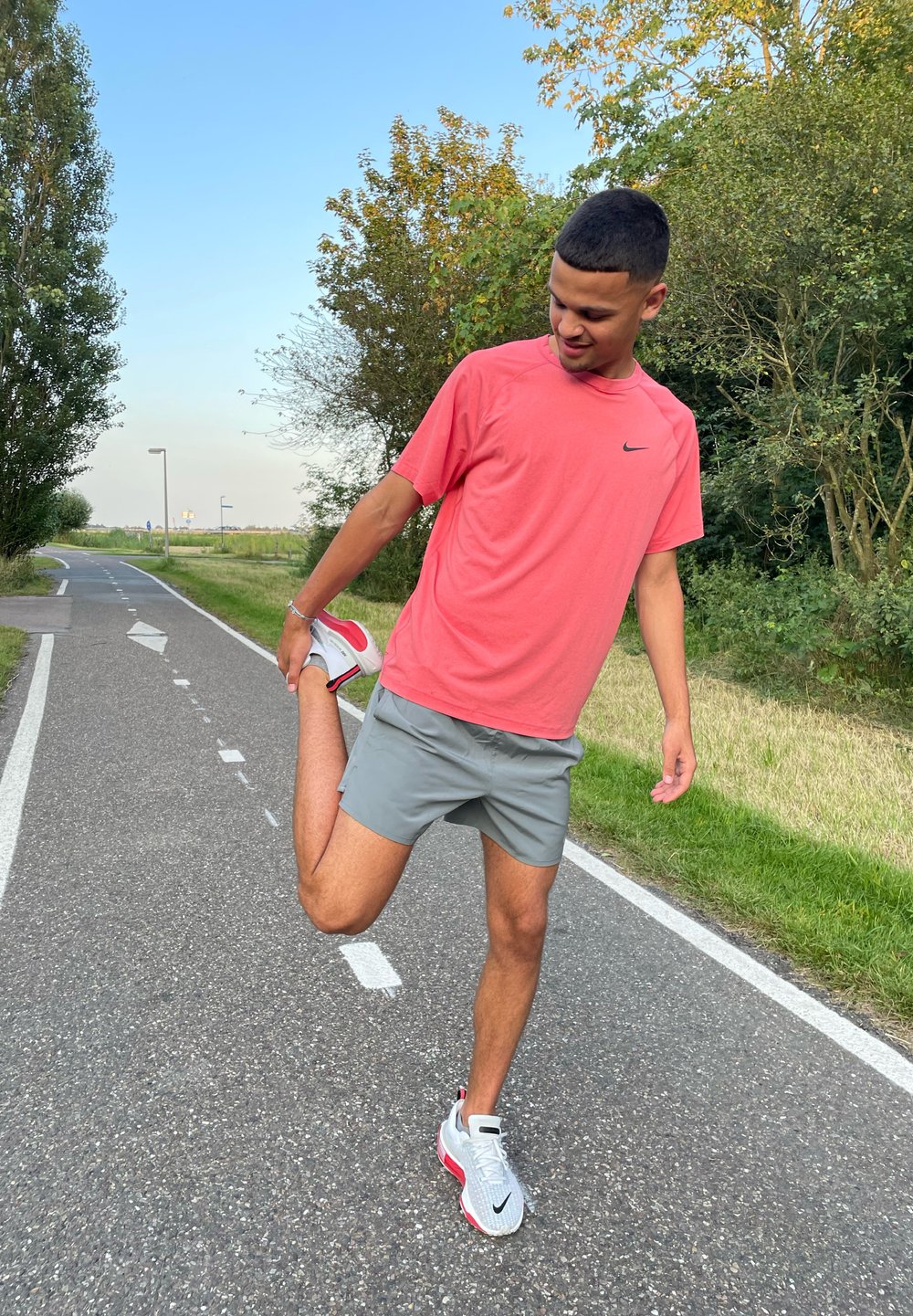 Nike rózsaszín rövidujjú póló és szürke rövidnadrág, amelyet egy személy visel, miközben nyújtja a lábát. Fehér és piros sportcipőt visel egy ösvényen.