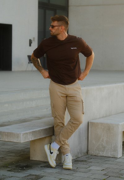 Camiseta marrón de cuello redondo, pantalones cargo beige con bolsillos laterales y zapatillas blancas con detalles en negro, de pie junto a un banco de concreto.