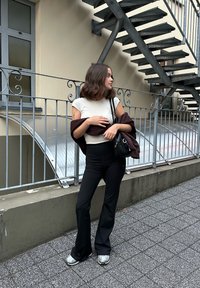 Fekete fodros nadrág, egy testhezálló fehér póló és egy sötét kabát van rajta. Egy kis fekete táska lóg a válláról, szemben egy acélszerkezettel.