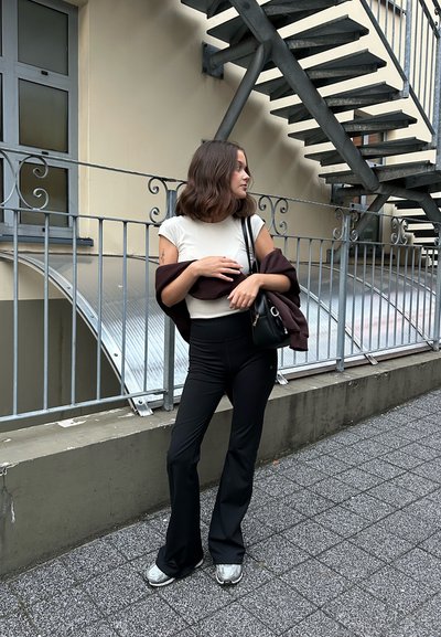 Llevan pantalones de campana negros, una camiseta ajustada blanca y una chaqueta oscura. De un hombro cuelga un pequeño bolso negro, apoyado contra una escalera de acero.