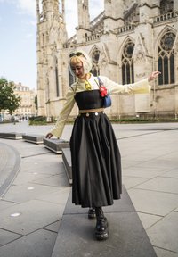 Persona equilibrándose en un banco de piedra, vistiendo un vestido negro, una camisa amarilla y botas, con una catedral gótica histórica de fondo.