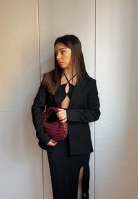 Vrouw met lang bruin haar, gekleed in een zwarte blazer en jurk met een split, die een geweven bordeauxrode handtas vasthoudt en tegen een textuurwand staat.