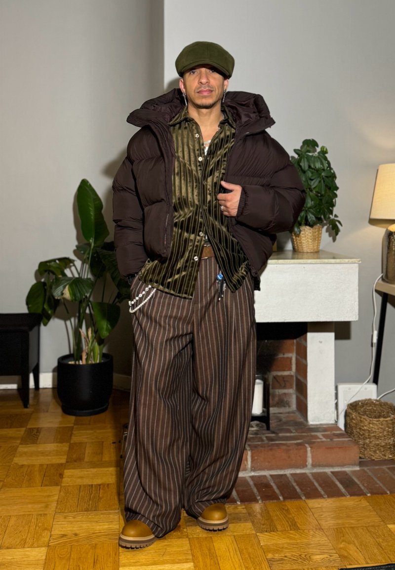 Homme à l'intérieur portant une veste matelassée marron, une chemise rayée olive, un pantalon large à fines rayures marron, des chaussures marron clair et une casquette plate verte, debout près de plantes et d'une cheminée.