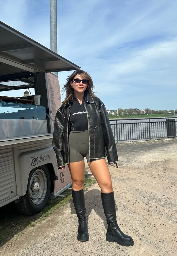 Junge Frau trägt Sonnenbrille, schwarze Lederjacke, schwarzes Top, olivfarbene Shorts und schwarze kniehohe Stiefel und steht in der Nähe eines Foodtrucks an der Uferpromenade.
