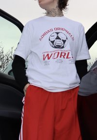 Hvit bomulls-T-skjorte med rødt grafisk trykk som viser en fotball og teksten «ADIDAS ORIGINALS WORLD», kombinert med løse røde shorts.