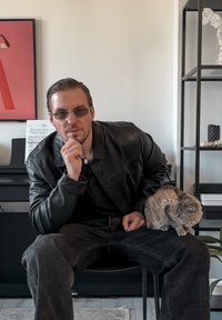 Blouson en cuir noir, chemise noire et jean foncé. Personne portant des lunettes de soleil, assise avec un chat gris sur les genoux. Partition musicale en arrière-plan.