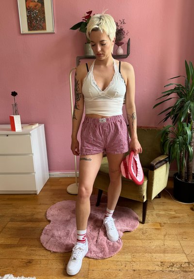 Top halter blanco con encaje, pantalones cortos a cuadros rojos y blancos, zapatillas blancas, diadema a rayas rojas y blancas, alfombra rosa, silla verde, planta.