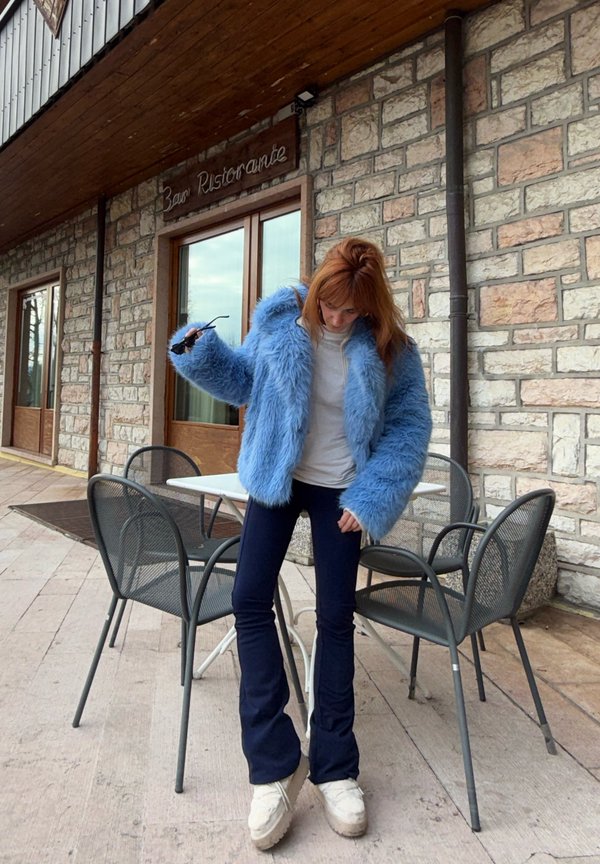 Person iført en blå, fnugget jakke, hvid skjorte, marineblå bukser og hvide sko, stående ved siden af et bord og metalstole udenfor en stenmuret bygning.