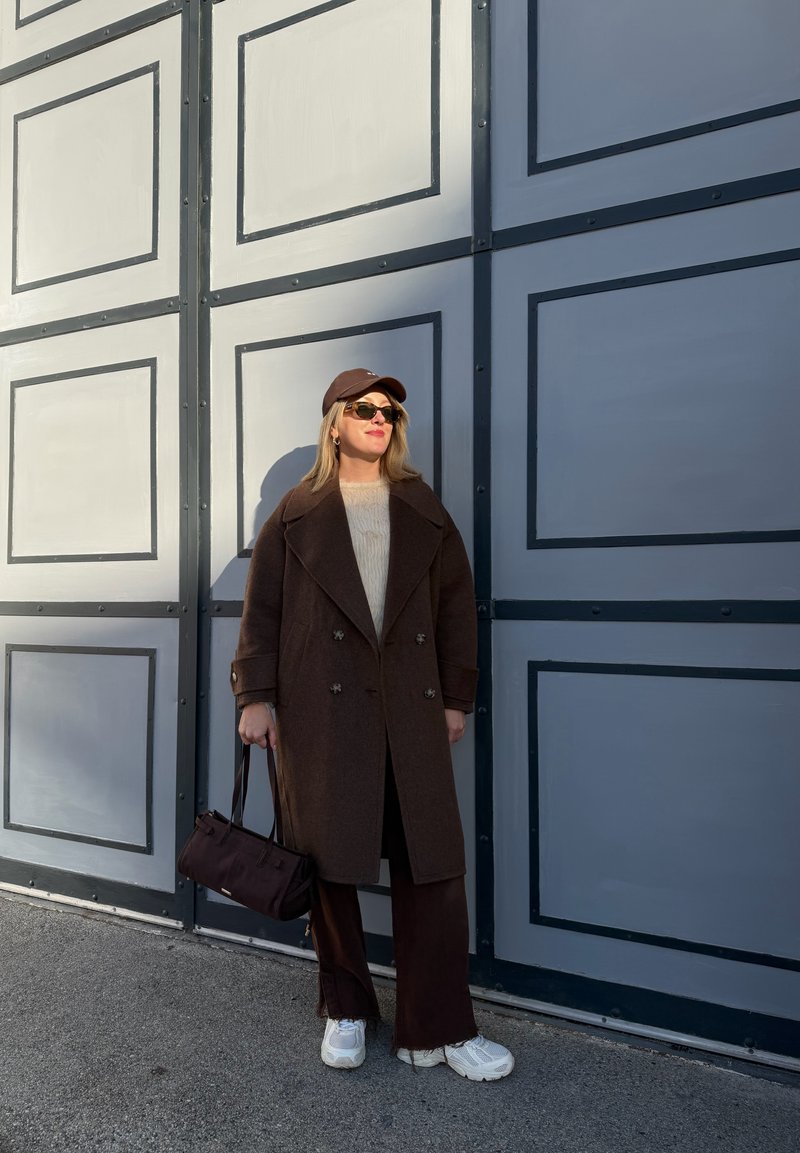 Manteau marron croisé sur un pull crème, pantalon foncé, baskets blanches et casquette foncée. Lunettes de soleil noires et petit sac à main.