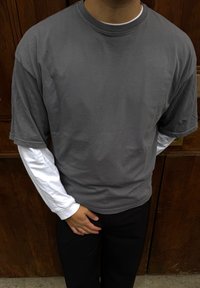 Personne portant un t-shirt à manches longues blanc superposé sous un t-shirt gris à manches courtes, un pantalon noir, debout avec une main reposant sur l'autre près d'une porte en bois.