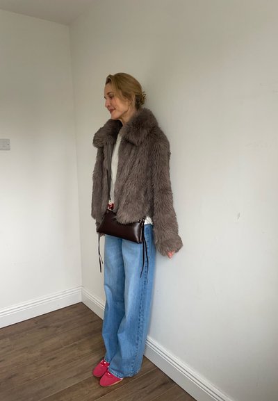 Mujer de pie en el interior con una chaqueta de piel sintética marrón, camiseta blanca, vaqueros anchos azules, zapatos rosas y un pequeño bolso bandolera marrón oscuro.