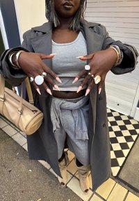 Persona ģērbta pelēkā mētelī, sportiskā topā, vaļīgās biksēs, bēšos zābakos un vairākās sudraba gredzenos un aprocēs, paceļ rokas, lai parādītu nagus.