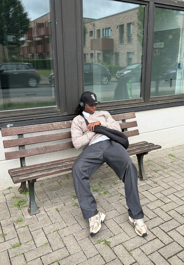 Un banco de madera presenta a una persona con un suéter beige y pantalones de rayas oscuros, que lleva una gorra negra y auriculares, sentada al aire libre.