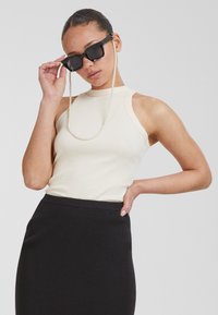 Beige bordás ujjatlan felső magas nyakkivágással, fekete bordás ceruzaszoknyával párosítva. Kiegészítők között fekete napszemüveg és gyöngy lánc található.