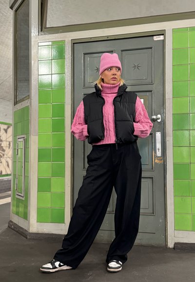 Chaleco acolchado negro sobre un suéter de punto trenzado rosa, combinado con pantalones negros holgados y zapatillas blancas con detalles negros, todo frente a azulejos verdes.
