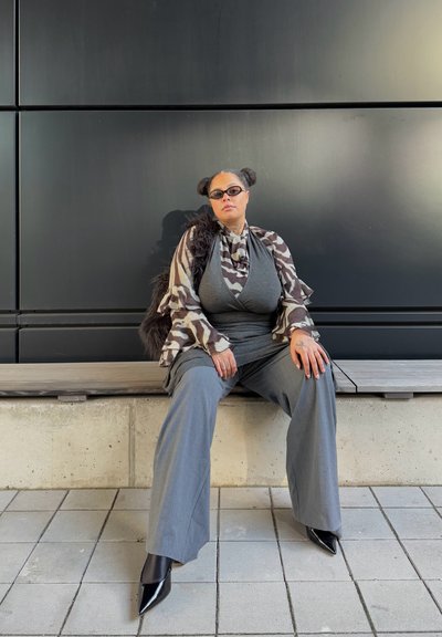 Pantalones de pierna ancha en gris combinados con una blusa gris. Una superposición fluida y con estampado cae sobre el conjunto. Los zapatos de tacón negro de punta y las gafas de sol completan el look.