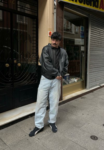 Chaqueta bomber de cuero negro sobre una camisa negra, combinada con jeans de pierna ancha de color azul claro y zapatillas deportivas negras, de pie sobre una acera.