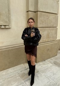 Giacca di pelle nera con maniche a sbuffo, gonna marrone con pizzo, e stivali neri al ginocchio. Tiene in mano una tazza di caffè. Sfondo semplice.