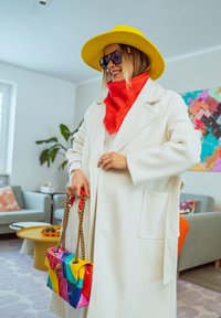 Femme portant un chapeau jaune, des lunettes de soleil noires, une écharpe rouge, un manteau blanc, tenant un sac à main rayé coloré à l'intérieur.