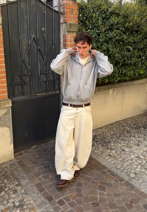 Joven ajusta la capucha de una sudadera gris con cremallera, lleva pantalones blancos anchos y zapatos marrones, está de pie en un camino adoquinado junto a una puerta metálica negra y arbustos verdes.