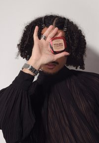Une personne avec des cheveux frisés et des pinces tient le fond de teint L'Oréal Infallible Fresh Wear devant un œil, portant un haut plissé noir et une montre en argent.