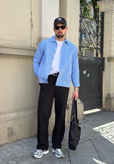 Hombre con gorra negra, gafas de sol, camisa azul a rayas, camiseta blanca, pantalones negros y zapatillas, está apoyado en una pared sosteniendo una bolsa tote negra.