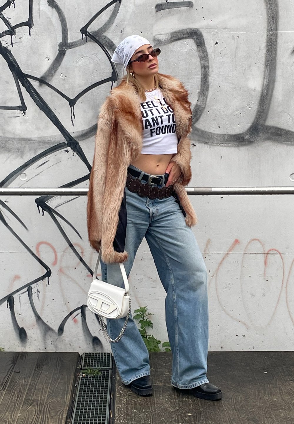 Model îmbrăcată cu un palton bej din blană sintetică, un tricou alb scurt cu text negru, blugi largi din denim, pantofi masivi negri și o geantă albă.