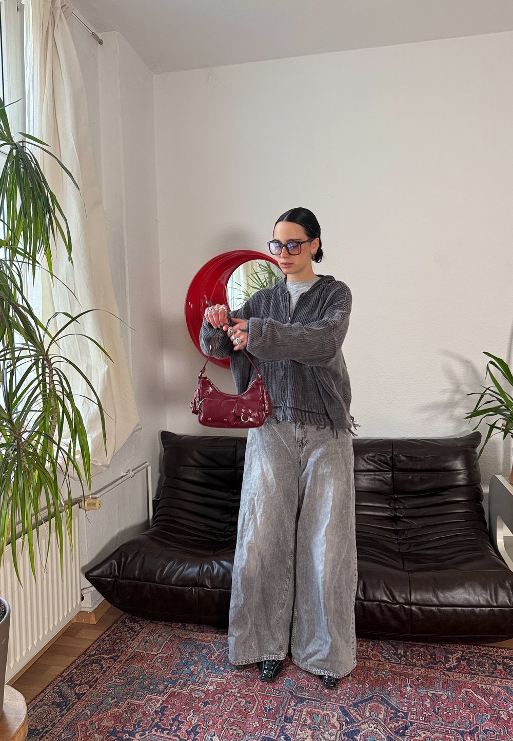 Veste zippée en velours côtelé gris, pantalons larges gris, et sac à main en cuir verni rouge. Debout devant un miroir rond rouge avec une plante verte à proximité.