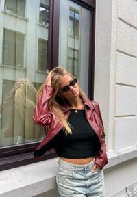 Červená kožená bunda, černý crop top a světle modré džíny. Minimalistické černé sluneční brýle. Odraz viditelný v okně za nimi.