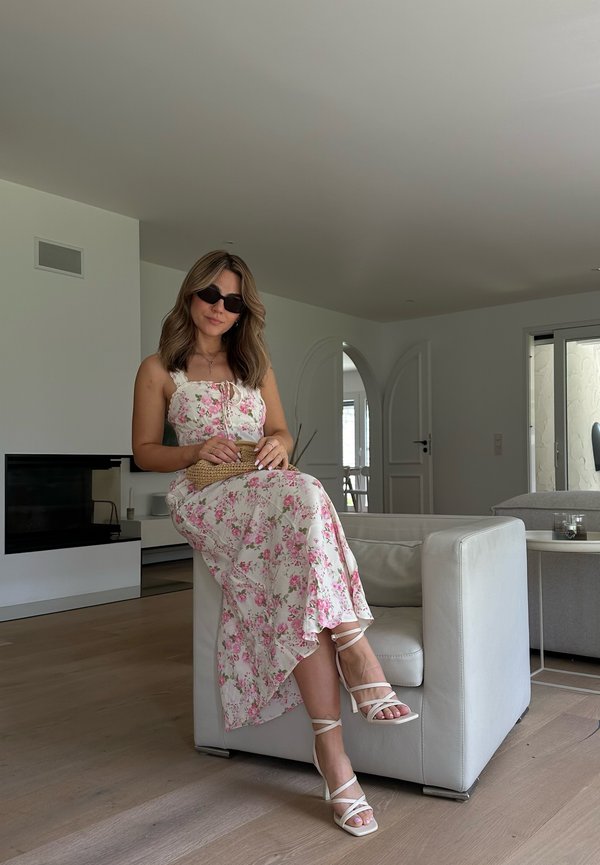 Abito floreale bianco con rose pink, caratterizzato da un corsetto aderente e una gonna fluida. Sandali con zeppa e cinturini incrociati. Interni neutri.