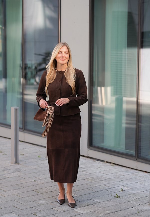 Femme aux longs cheveux blonds portant une veste et une jupe marron, tenant un petit sac à main marron et une écharpe, debout sur un trottoir près de fenêtres en verre.