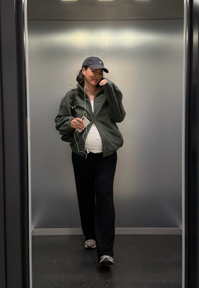 Mujer embarazada caminando en un ascensor, vestida con chaqueta oscura, pantalones negros, gorra, auriculares, sosteniendo un teléfono inteligente y una pequeña bolsa negra.