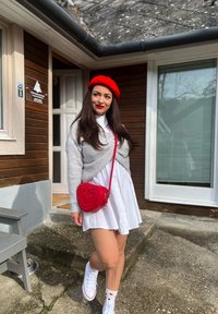 Röd hjärtformad crossbody-väska, grå stickad sjal, vit oversized skjortklänning, vita sneakers och strumpor med hjärtmönster. Bakgrund av trä och glas.