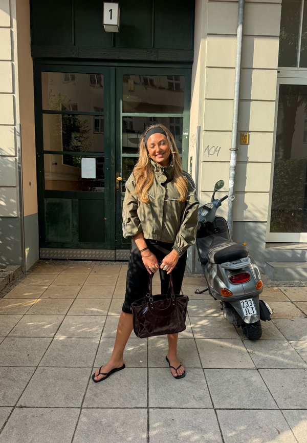 Femme en veste verte et short noir tenant un sac, debout sur un trottoir près d'un scooter moteur argenté garé à l'entrée d'un bâtiment avec des portes vertes.