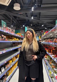 Cappotto nero, maglietta grigia e pantaloni corti neri con capelli biondi in un corridoio di vendita. Scaffali riforniti con vari prodotti snack, alcune confezioni colorate sono visibili.