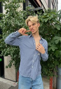 Mladý člověk s blond vlasy, oblečený v pruhovaném tričku a džínách, pózuje s prstem na tváři, mžourá a stojí vedle zelených listnatých rostlin venku.