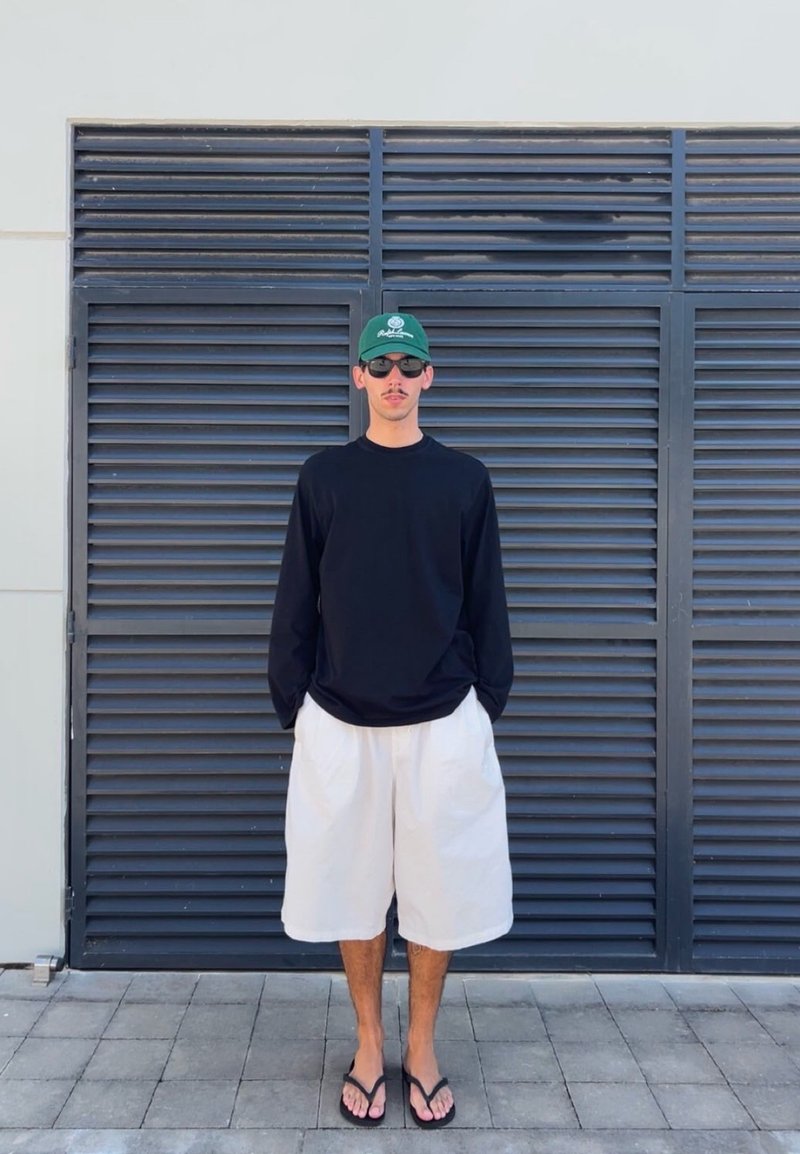 Homme portant une chemise noire à manches longues, un short blanc, une casquette verte, des lunettes de soleil et des tongs debout devant une porte à persiennes gris foncé.