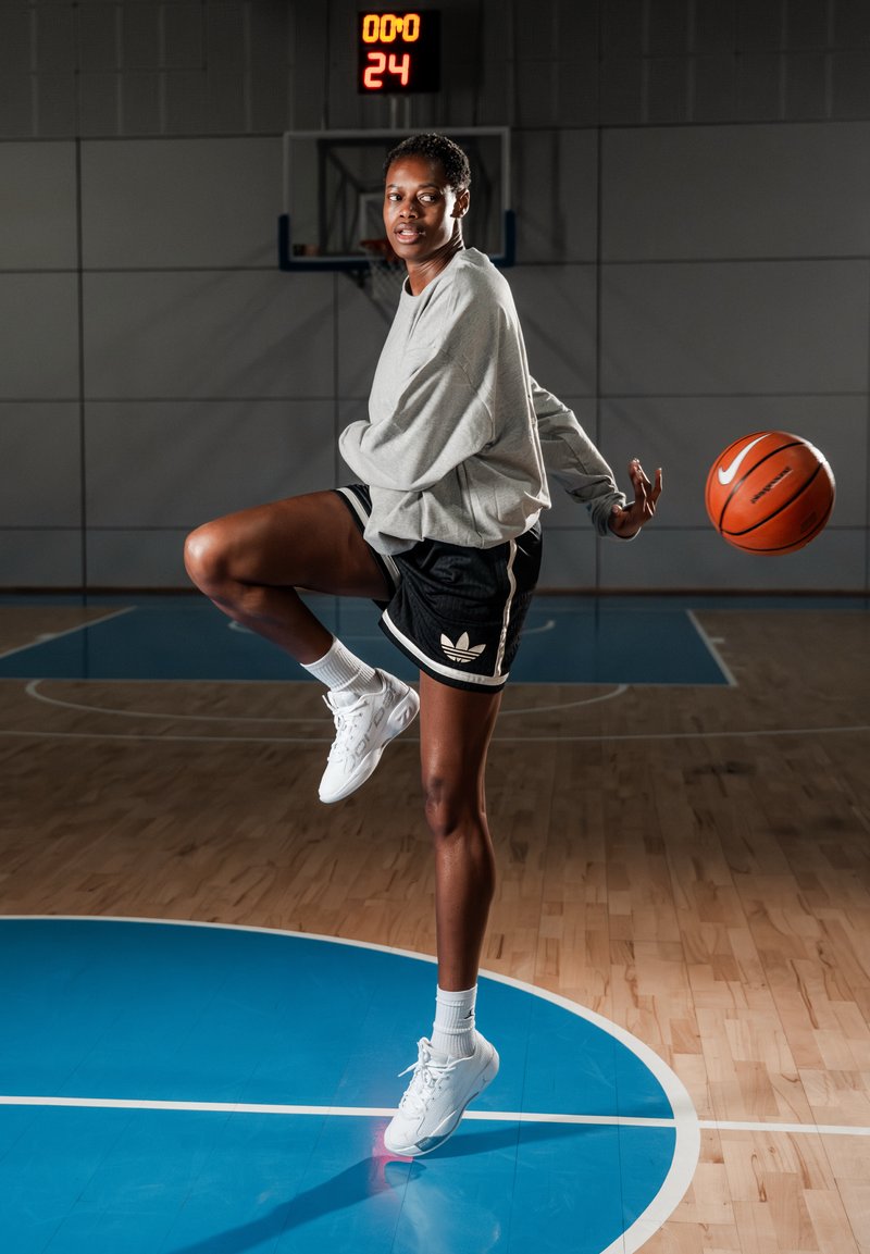 Giocatrice di basket in felpa grigia e pantaloncini neri mentre dribbla a metà campo, con il tabellone che mostra 0:00 e 24 secondi.