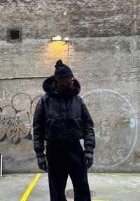 Fekete pehelydzseki szőrme szegéllyel, kapucnis kialakítással és cipzáras zsebekkel. Fekete kesztyűvel és bordázott sapkával viselve, texturált fal előtt.