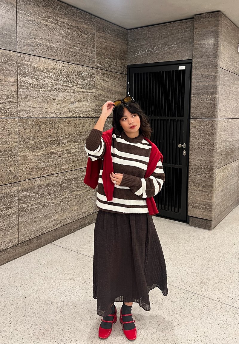 Brown and white striped sweater layered with a red cardigan, paired with a long black textured skirt and red shoes with straps.