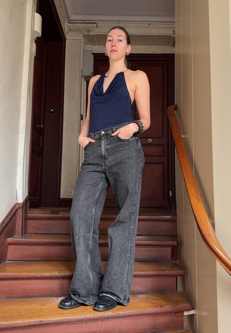 Haut dos nu bleu marine, jean en denim noir à jambes larges, bracelet argenté, chaussures noires, debout sur des escaliers en bois devant un mur miroir.