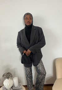 Sort skræddersyet blazer over en sort tekstureret top, kombineret med sølvfarvede palietbukser. Holder en lille sort taske, med en hvid væg i baggrunden.