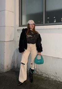 Νεαρή γυναίκα που φοράει μπεζ καπέλο, μαύρο πανωφόρι από ψεύτικη γούνα, τοπ καμουφλάζ, φαρδιά κρεμ παντελόνια με σκισίματα, μαύρες μπότες, κρατώντας τυρκουάζ τσάντα δίπλα σε λευκό τοίχο.