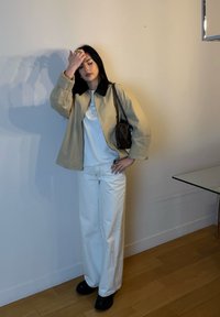 Giacca beige con colletto nero, camicia bianca e pantaloni bianchi larghi. Stivali neri e una piccola borsa marrone. Interno minimalista.