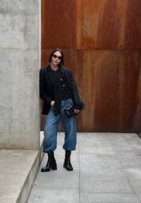 Blazer preto, top preto franzido, jeans azul de perna larga, botas pretas até o joelho, mala preta. Texturas suaves com detalhes de tecido em camadas.