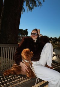 Femme portant des lunettes de soleil et une veste sombre assise sur un banc en plein air, tenant sur ses genoux un petit chien marron vêtu d'un pull.