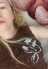 Femeie blondă întinsă pe o suprafață cu perne roșii texturate, purtând un top maron cu o broșă argintie în formă de floare.