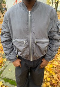 Sivá bomber bunda s textúrovaným povrchom, vybavená vreckami na zips vpredu, rebrovanými manžetami a centrálnym zapínaním na zips. Ľahký dizajn.