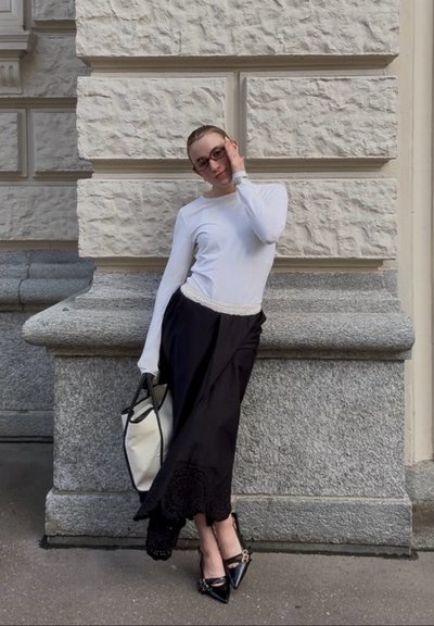Mujer con gafas, camiseta blanca de manga larga, falda negra, zapatos negros puntiagudos, sosteniendo un gran bolso tote blanco, apoyada contra una pared de piedra.