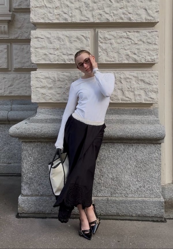 Γυναίκα με γυαλιά, λευκό μπλουζάκι μακρυμάνικο, μαύρη φούστα, μαύρα μυτερά παπούτσια, κρατώντας μια μεγάλη λευκή τσάντα, στηρίζεται σε έναν πέτρινο τοίχο.